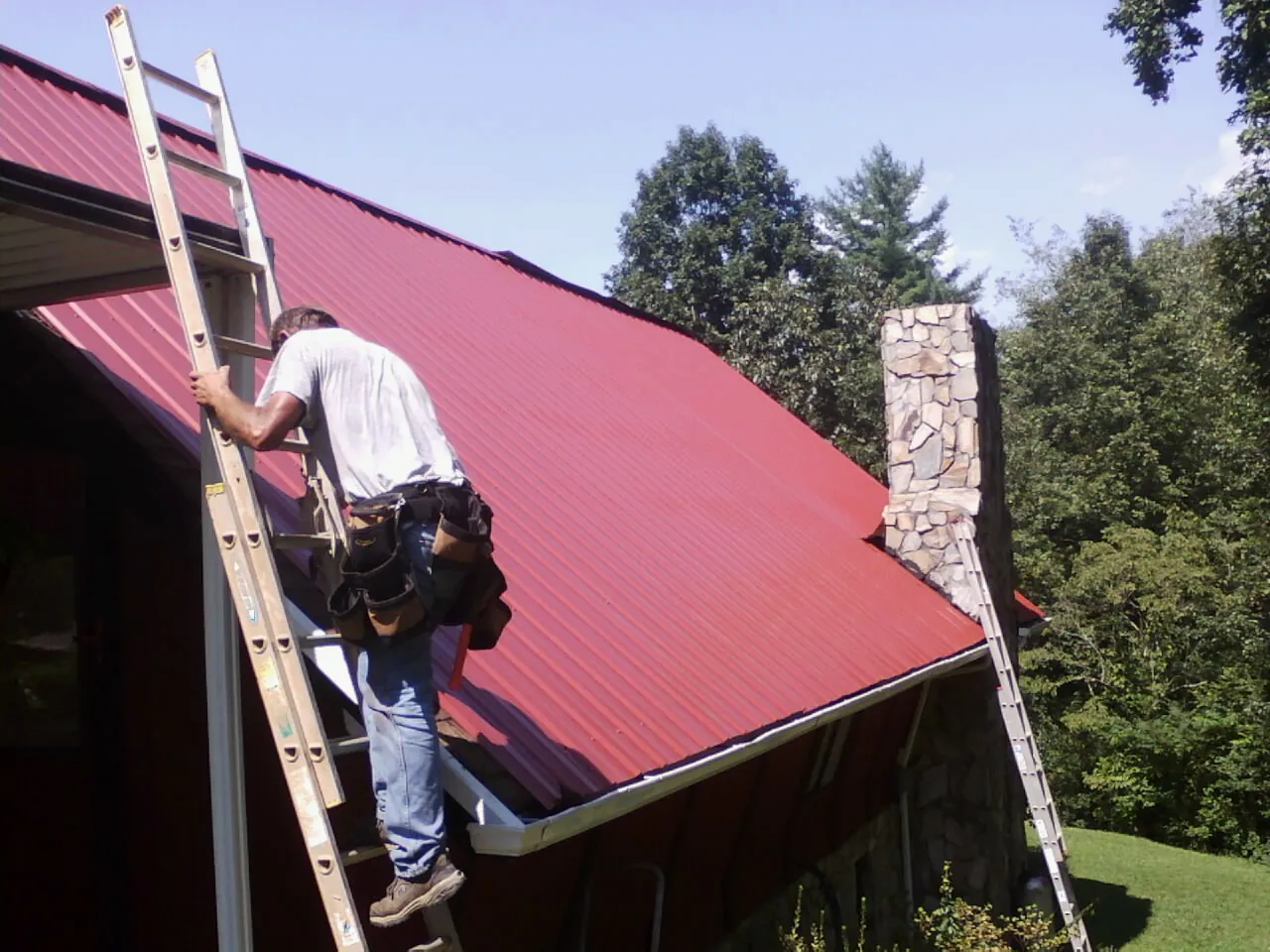 Roofer on a ladder beside a red metal roof and stone chimney — Soffit & Fascia Repair by Roofing Brothers in Bogart